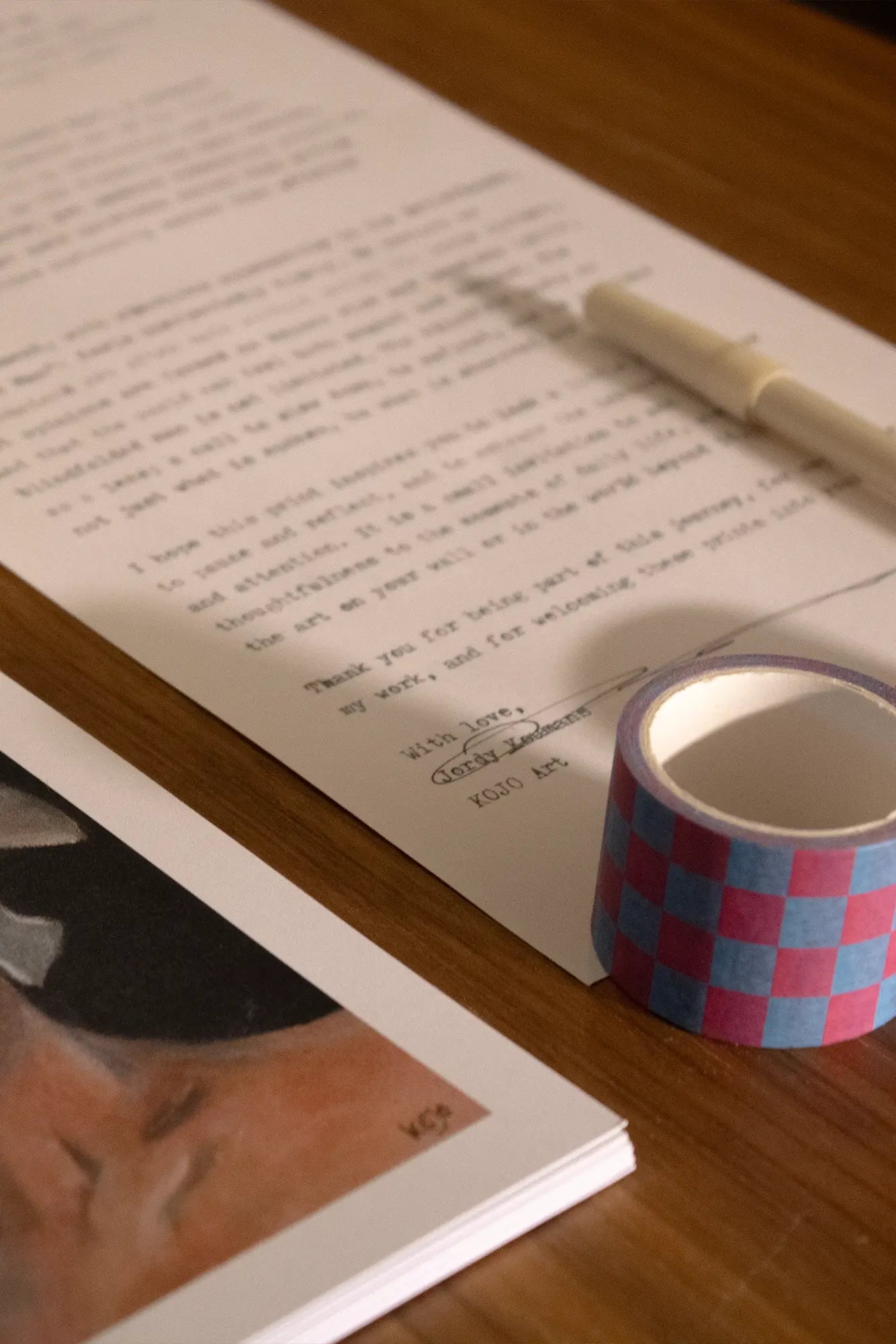 Een handgeschreven brief van KOJO Art op een houten tafel, met een pen, kleurrijke washi tapes en een art print gedeeltelijk zichtbaar naast de brief, klaar om via snailmail te worden verzonden.