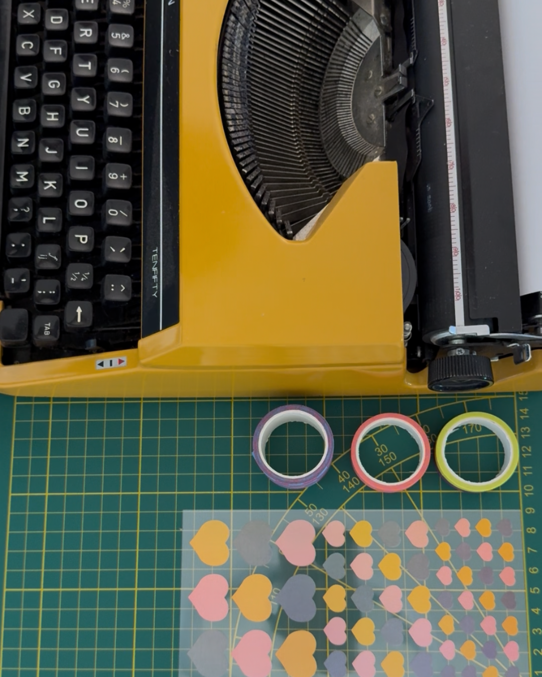 We zien een typemachine op een wormpad met daarnaast washi tape en stickers in de vorm van een hartje. Dit alles is onderdeel van de snail mail art print club die KOJO Art elke maand verstuurt.